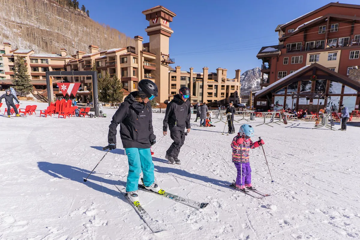 A joyful winter scene showing a family skiing together, weaving through snowy slopes with resort buildings in the background. The landscape is blanketed in fresh snow, capturing the thrill and togetherness of a day on the mountain.