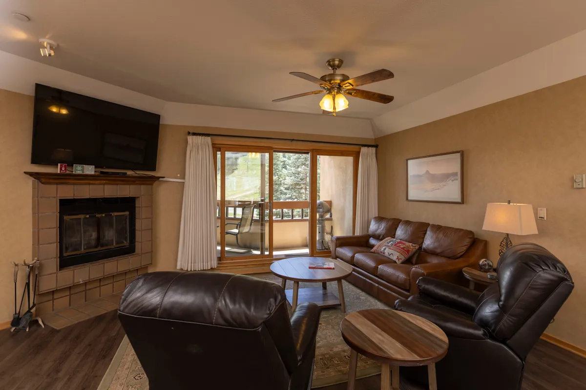 A cozy living area featuring a prominent wood-burning fireplace, a plush sofa, and a wooden coffee table. Large windows take up most of one wall, framing expansive views of the Needles Mountain Range and allowing natural light to pour into the space. The overall atmosphere is warm and inviting, ideal for relaxing after a day on the slopes.