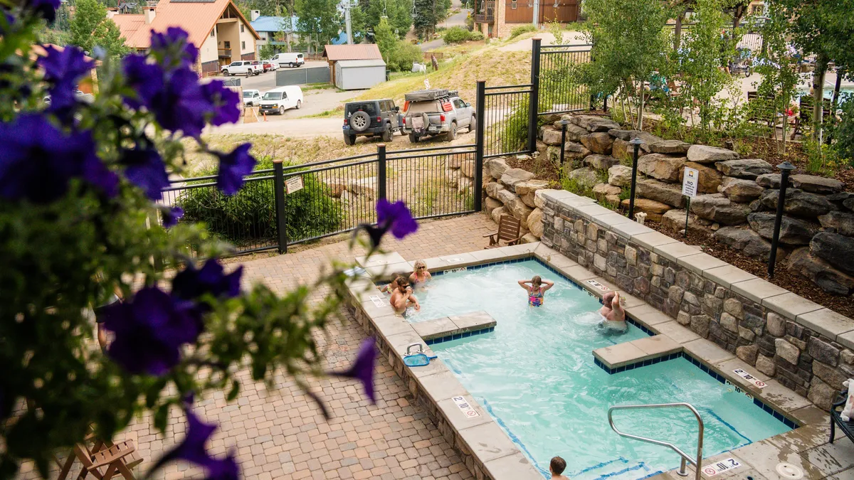 A sunlit outdoor pool surrounded by flowering plants and cozy patio seating. The inviting water and relaxed seating area create an atmosphere of leisure and summer indulgence.