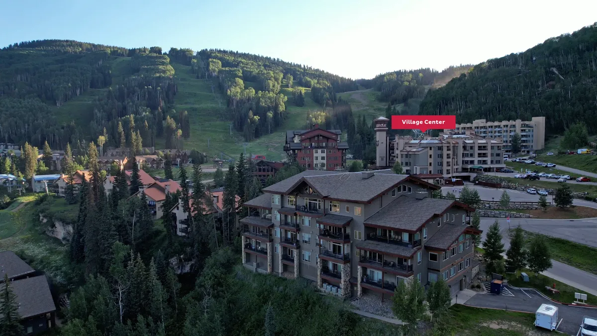 A vibrant exterior view capturing the mountain landscape, residential resort buildings, and a sign marking the Village Center. People can be seen exploring the area, suggestive of seasonal festivals or community events, with the majestic mountains serving as a dramatic backdrop.