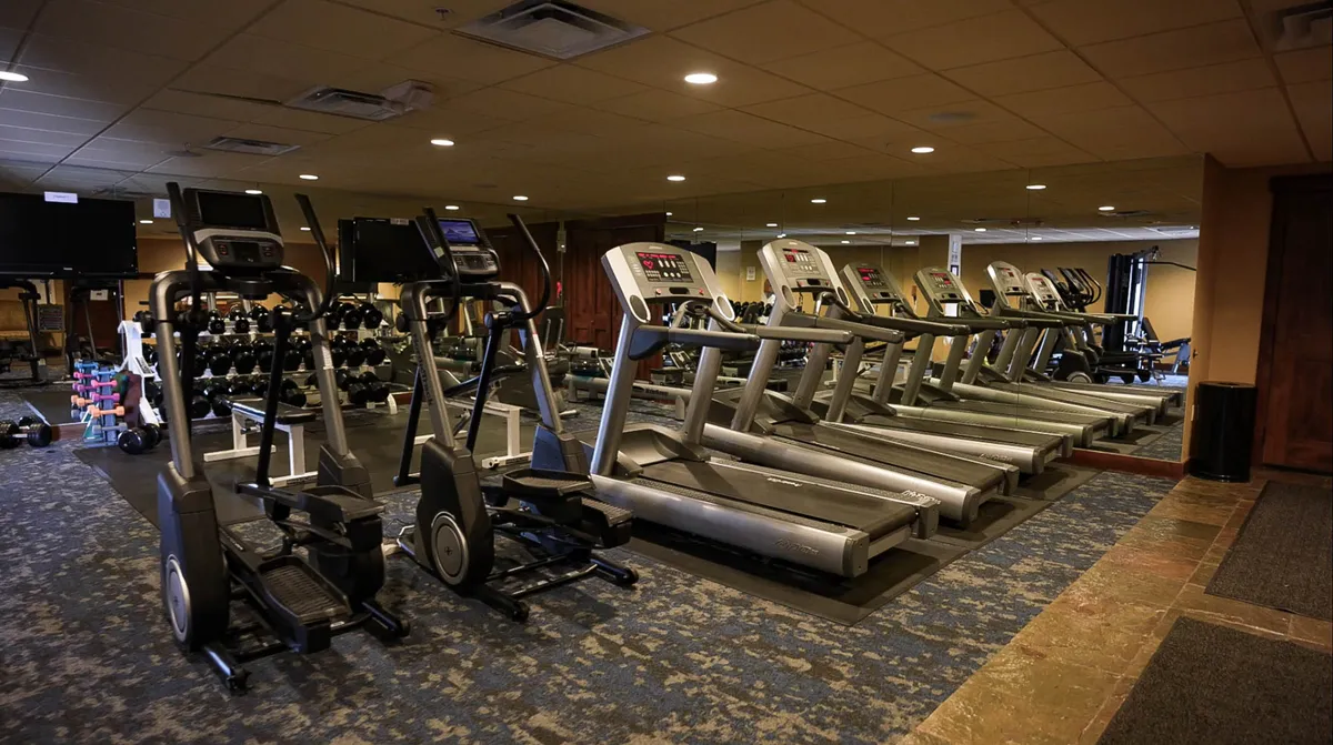 A modern, well-equipped fitness center filled with treadmills, elliptical machines, dumbbells, and a mirrored wall. The bright, airy space is designed for energetic workouts and personal wellness routines.