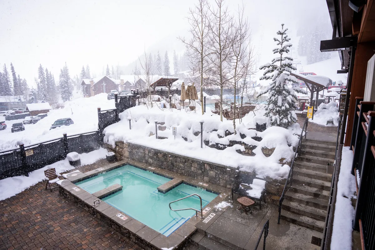 An exterior scene featuring a hot tub surrounded by snow-covered trees and mountains, with steam rising from the water. The tub is set amidst this picturesque alpine landscape, evoking relaxation and luxury after winter sports.