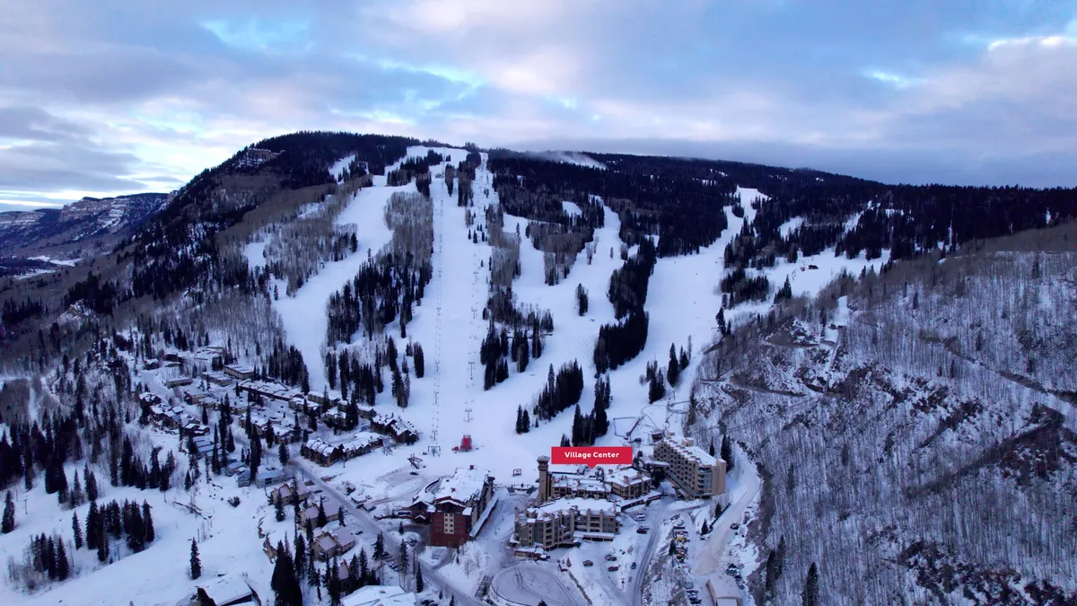 A sweeping exterior view of the ski resort with snow-covered ski slopes running down to a lively village center, surrounded by majestic mountains and frosted trees. The scene captures skiers and snowboarders in action, with the resort buildings nestled at the base and the soft glow of sunrise illuminating the landscape.