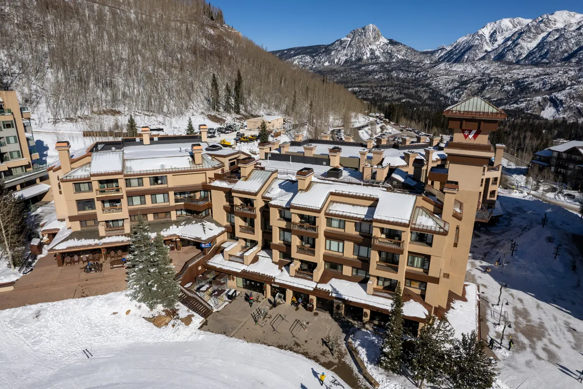 A picturesque exterior view displaying snow-covered rooftops with a dramatic mountain backdrop, outdoor seating areas, and a ski lift visible in the distance. The scene evokes a lively resort atmosphere, with hints of both winter adventure and tranquil relaxation.