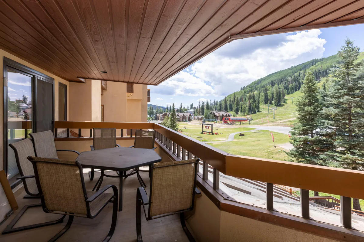 The image showcases an inviting balcony with comfortable seating and a table, overlooking breathtaking, panoramic mountain views at sunset. The tranquil mountain landscape invites relaxation and socializing, while the balcony setting suggests the perfect spot for sipping drinks and soaking in the scenery after a day on the slopes.