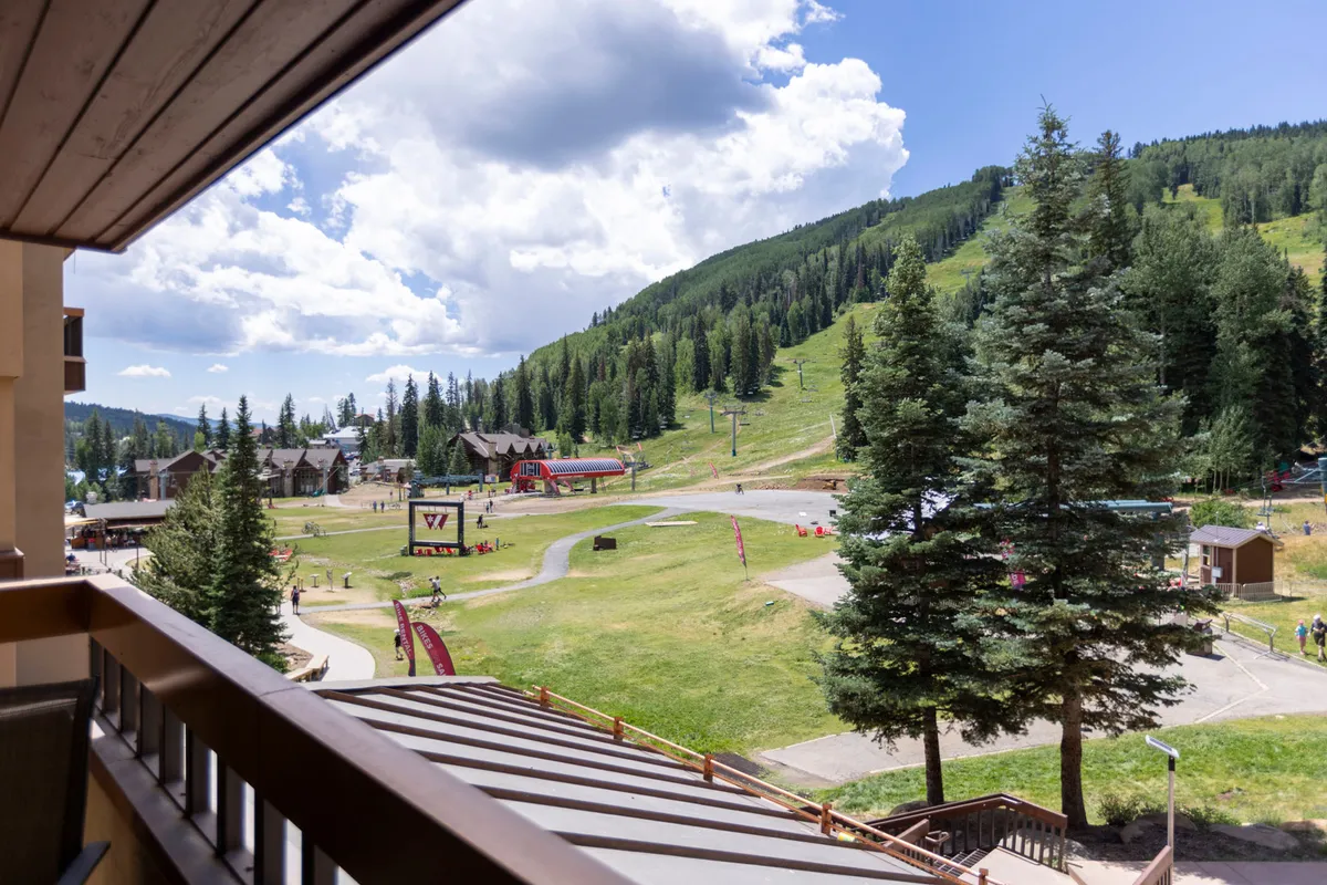 An expansive mountain landscape with ski lifts ascending green and snow-dusted slopes, framed by wooden resort architecture. The scene evokes adventure, with visible ski runs and a hint of balcony views that promise dramatic sunsets and a front-row seat to mountain action.
